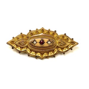 May include: A gold brooch with an oval center featuring a red gemstone surrounded by two white pearls. The brooch has a detailed, ornate design.