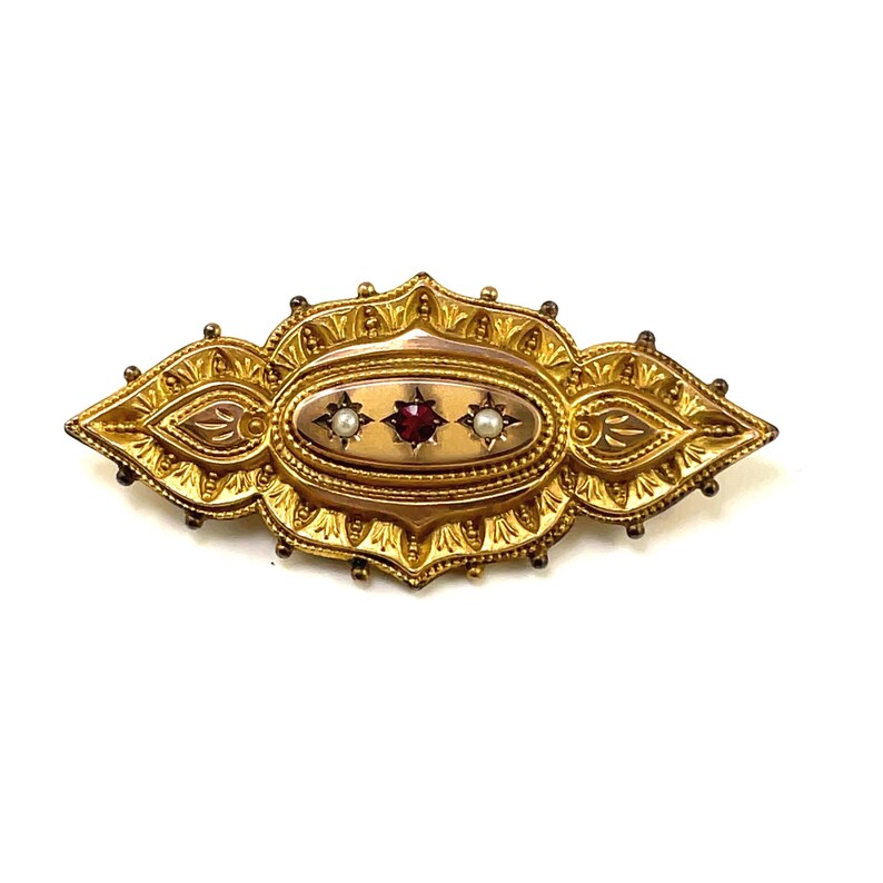 May include: A gold brooch with an oval center featuring a red gemstone surrounded by two white pearls. The brooch has a detailed, ornate design.