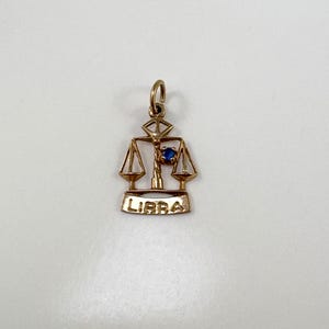 May include: Gold-coloured Libra charm with a blue gemstone centre. The charm features the scales of justice design, with the word "LIBRA" inscribed below. A small loop is at the top for attachment to a necklace or bracelet.