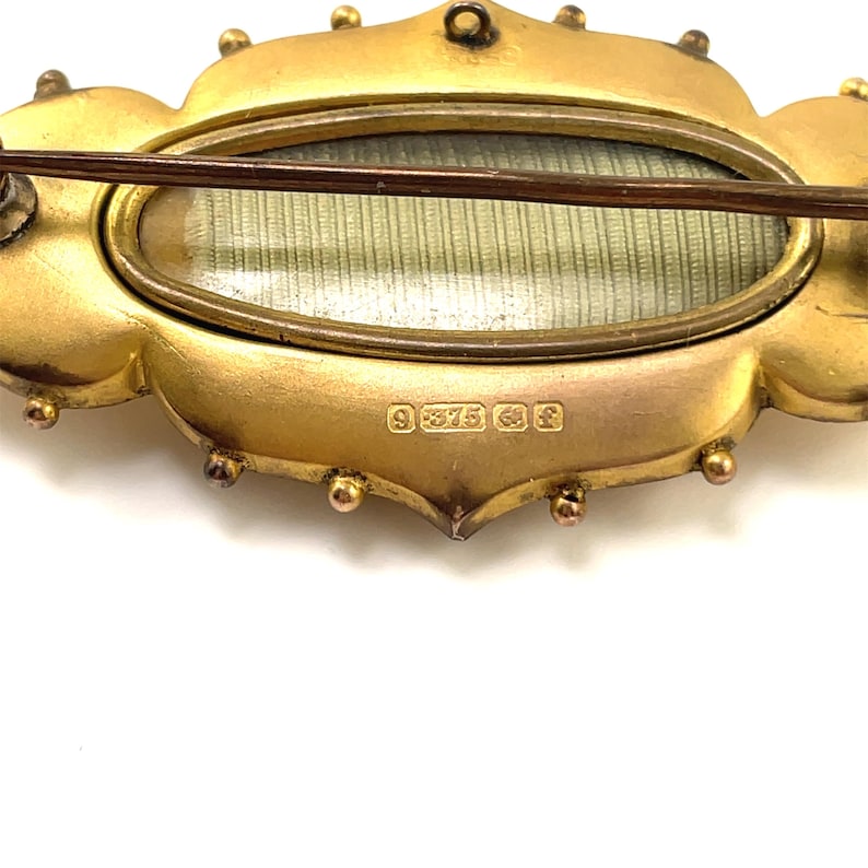 May include: Gold-toned oval locket brooch with a glass front and a back that is engraved with the hallmark "9 375 38 F".