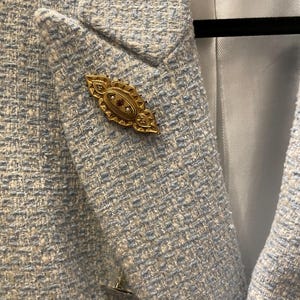 May include: A light blue blazer with a white and light blue patterned design. A gold brooch with red and white stones is pinned to the lapel.