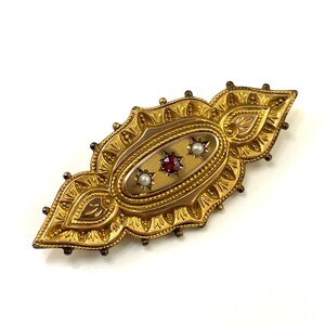 May include: A gold brooch with an oval-shaped center featuring a red gemstone and two white pearls. The brooch is intricately detailed with a floral design.