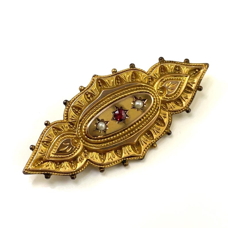 May include: A gold brooch with an oval-shaped center featuring a red gemstone and two white pearls. The brooch is intricately detailed with a floral design.