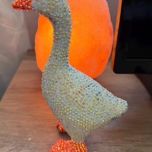 May include: A decorative goose figurine completely covered in iridescent white crystals. The beak and webbed feet are accented with bright orange crystals. The goose has a single small black bead for an eye and stands in a profile position.