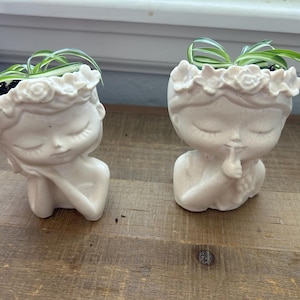 May include: Two off-white ceramic planters shaped like serene figures with closed eyes and floral headbands. Each planter holds a small green plant. One figure rests its chin on its hand, while the other holds a finger to its lips.