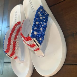 May include: Pair of white flip-flops with beaded straps. One strap features red and white beads, the other blue and white beads. The word "ROCKET" is printed on the side of each shoe.