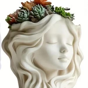 May include: A white ceramic planter shaped like a woman's face with closed eyes and flowing hair. The top of the planter is filled with a variety of succulents in shades of green, orange, and red. A decorative item for home decor.