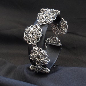 May include: A silver bracelet with a black leather band and multiple chainmail flower designs.