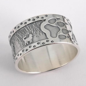 Può includere: Anello in argento con un design dettagliato raffigurante la sagoma di un cane, impronte di zampe ed erba alta. L'anello ha una fascia larga e una finitura lucida.