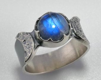 Könnte beinhalten: Ein Silberring mit einem runden, schillernden blauen Mondstein-Cabochon in einer gewellten Fassung. Die Ringschiene weist an den Seiten eine texturierte, gehämmerte Oberfläche auf, die beidseitig des zentralen Edelsteins eine Mondsichelform bildet.
