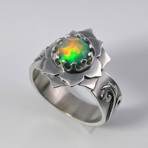 May include: A silver ring with a green and orange opal gemstone set in a flower-shaped design. The band is decorated with a swirling pattern.