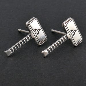 Pode incluir: Um par de brincos de prata em forma de martelo de Thor, Mjolnir. Cada martelo apresenta uma cabeça retangular com um desenho de nó celta e uma pega detalhada. Os brincos são exibidos numa superfície cinzenta escura.