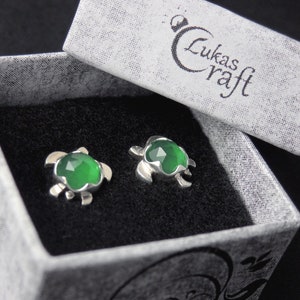 Può includere: Un paio di orecchini a forma di tartaruga in argento con accenti di pietre preziose verdi, presentati in una scatola bianca con il testo "Lukas Craft" sul coperchio.