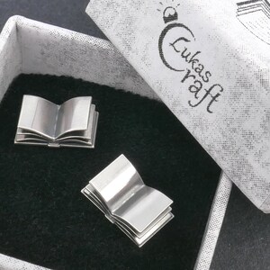 May include: Two silver book-shaped cufflinks in a white box with the text "Lukas Kraft" on the lid. The cufflinks are open and have a detailed design.