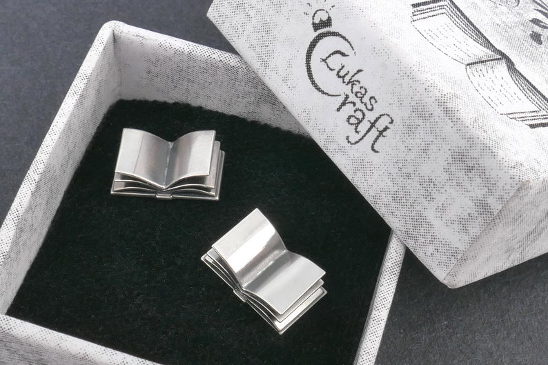 May include: Two silver book-shaped cufflinks in a white box with the text "Lukas Kraft" on the lid. The cufflinks are open and have a detailed design.