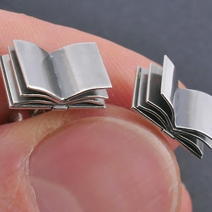 May include: A pair of silver book cufflinks. The cufflinks are shaped like open books with multiple pages.