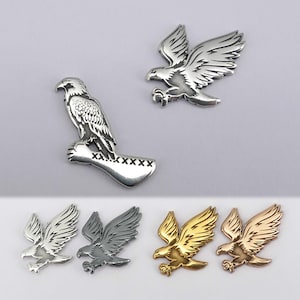 Puede incluir: Broches de metal con forma de águila en plata, gris, oro y oro rosa. Un águila está posada en una rama, mientras que otras están en vuelo. Los broches tienen diseños detallados de plumas y se muestran sobre un fondo gris claro.