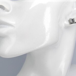 May include: Silver book-shaped stud earrings. The earrings are small and delicate, and they are shown on a white mannequin's ear.