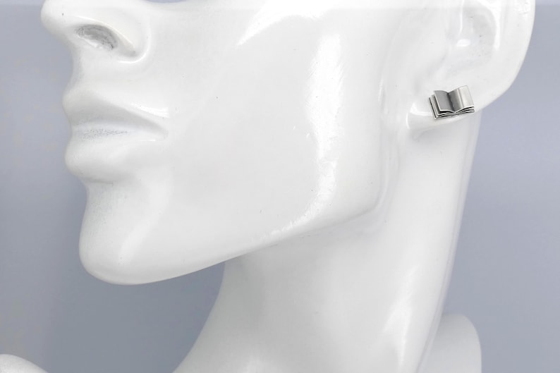 May include: Silver book-shaped stud earrings. The earrings are small and delicate, and they are shown on a white mannequin's ear.