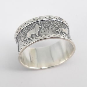 Può includere: Un anello largo color argento con un design dettagliato. L'anello presenta un rilievo di due cani in un campo erboso, con impronte di zampe lungo i bordi superiore e inferiore. L'anello ha un interno lucido e brillante.