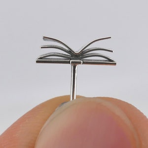 May include: A silver book charm with pages that are open. The charm is attached to a post that is designed to be worn as a piercing.