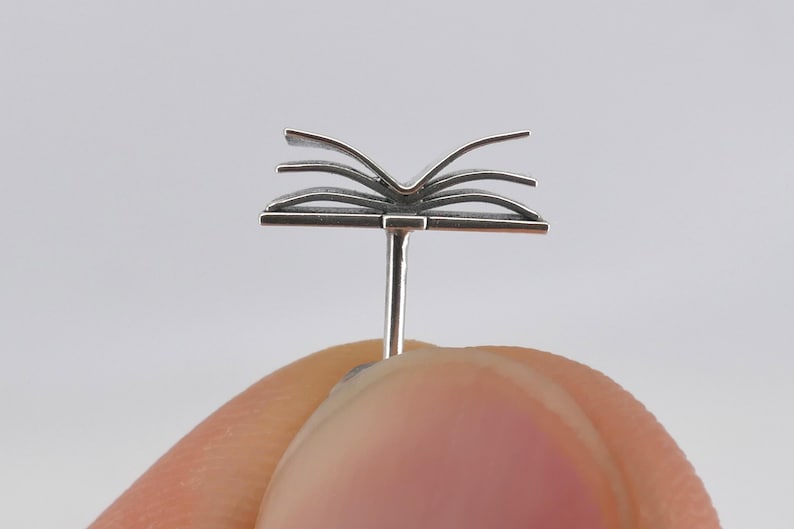 May include: A silver book charm with pages that are open. The charm is attached to a post that is designed to be worn as a piercing.