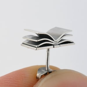 May include: Silver book-shaped stud earring. The earring is made of three layers of metal that are shaped like an open book. The earring is attached to a post that goes through the ear.