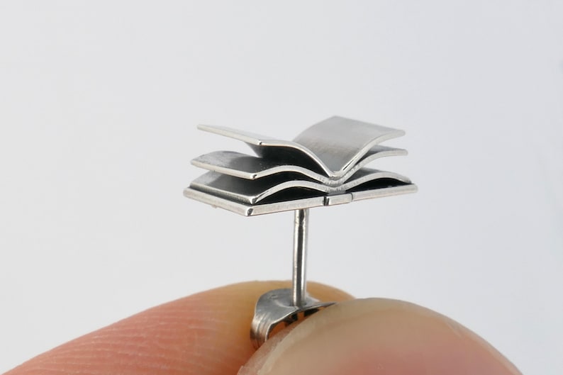 May include: Silver book-shaped stud earring. The earring is made of three layers of metal that are shaped like an open book. The earring is attached to a post that goes through the ear.