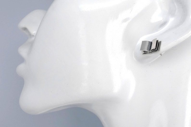 May include: A silver book-shaped stud earring. The earring is shown on a white mannequin head.