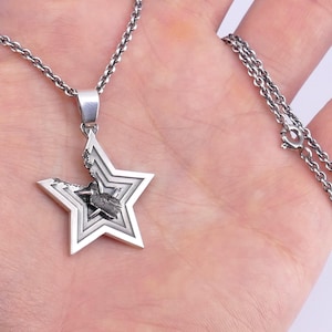 May include: A silver chain necklace with a star-shaped pendant. The star is made of multiple layers and has a small, dark gray stone in the center.