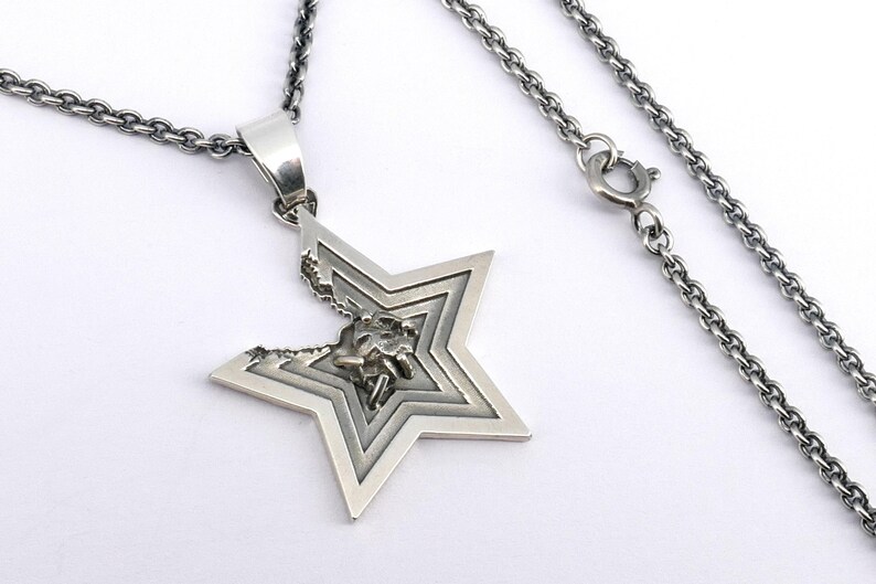 May include: A silver chain necklace with a star-shaped pendant. The star is made of multiple layers of metal, creating a three-dimensional effect. The pendant has a small, intricate design in the center.