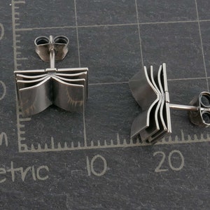 May include: A pair of silver book-shaped stud earrings. The earrings are made of metal and have a detailed design of a book with multiple pages.
