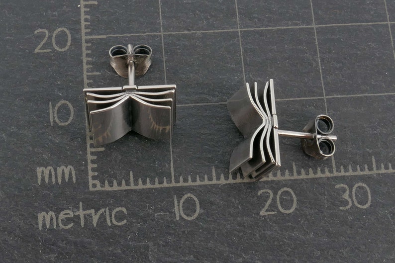 May include: A pair of silver book-shaped stud earrings. The earrings are made of metal and have a detailed design of a book with multiple pages.