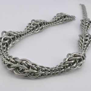 Op de afbeelding: Een zilveren ketting met een uniek, ingewikkeld ontwerp. De ketting is gemaakt van kleine, met elkaar verbonden metalen ringen die een reeks ronde, getextureerde vormen vormen.