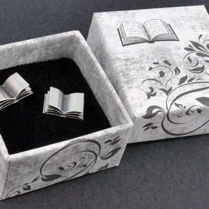 May include: A pair of silver book-shaped cufflinks in a grey and black patterned box with a black velvet interior. The box lid features a black and white illustration of an open book with a floral design.