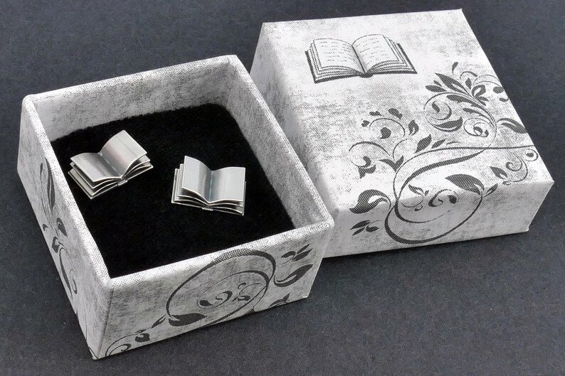 May include: A pair of silver book-shaped cufflinks in a grey and black patterned box with a black velvet interior. The box lid features a black and white illustration of an open book with a floral design.