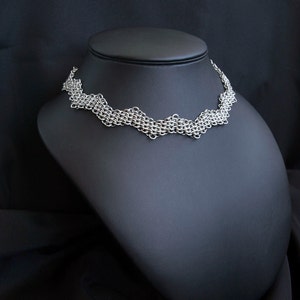 May include: A silver chainmail necklace with a delicate, wave-like design. The necklace is displayed on a black mannequin bust.