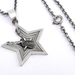 May include: A silver chain necklace with a star-shaped pendant. The star is made of layered metal and has a small black stone in the center.