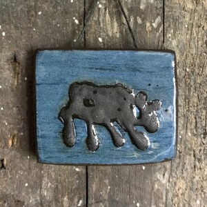 Può includere: Piccola decorazione murale rettangolare in ceramica con un design stilizzato di una mucca. La mucca è grigio scuro con macchie nere, su sfondo blu. Il pezzo è appeso con uno spago ed è montato su una superficie di legno invecchiato.