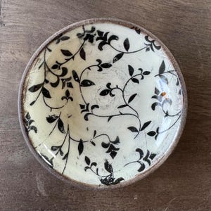 Puede incluir: Un pequeño cuenco de cerámica blanca con un patrón floral negro. El cuenco tiene un exterior ligeramente rugoso y texturizado.