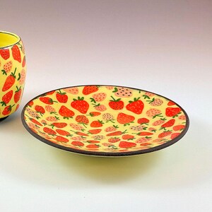 May include: A ceramic plate featuring a yellow backdrop adorned with a pattern of red and pink strawberries. The plate has a dark brown rim. A matching yellow cup with a strawberry pattern is partially visible.