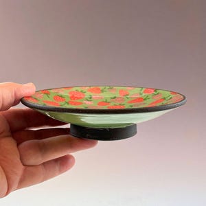 May include: A small, round ceramic dish with a green base and a black rim. The dish is decorated with a pattern of red strawberries on a green background. The dish is held by a hand.