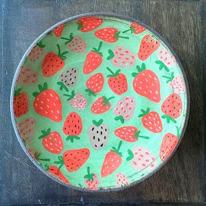 Könnte beinhalten: Eine runde, flache Schale mit hellgrünem Innenraum und einem Muster aus roten und weißen Erdbeeren mit grünen Stielen. Der Rand der Schale ist dunkelgrau. Die Erdbeeren sind unterschiedlich groß und über die Oberfläche verteilt.