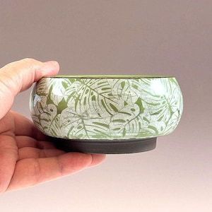 May include: A round ceramic bowl featuring a light green base color with a repeating white monstera leaf pattern. The bowl has a smooth, glossy finish on the upper portion and a contrasting matte dark charcoal gray base at the bottom.