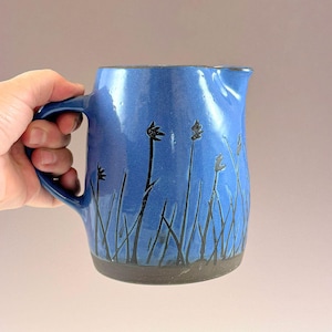 Op de afbeelding: Een blauwe keramische kan met een donkergrijze voet en een handvat. De buitenkant is voorzien van een zwart sgraffito-ontwerp van lange, slanke grassen of rietstengels die vanaf de onderkant omhoog lopen op de kan met een kleine schenktuit.