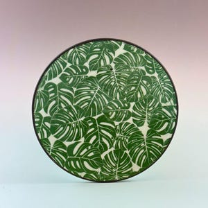 May include: Round decorative tray featuring a repeating pattern of green monstera leaves on a white background. The tray has a dark brown rim. The design is a botanical theme, suitable for home decor.