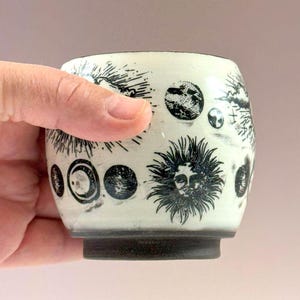 May include: A ceramic cup with a cream-colored glaze featuring black ink illustrations of celestial bodies including a sun with a face, various moon phases, and stars. The cup has a dark, unglazed base and a rounded, handleless cylindrical shape.