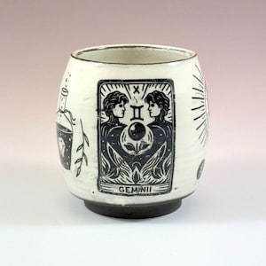 Op de afbeelding: Een witte keramische beker met een donkerbruine voet en zwarte inktillustraties. Het centrale ontwerp is een rechthoekige tarotkaartafbeelding met het label GEMINI, twee figuren en een bol. Links staat een fles en rechts een zon.