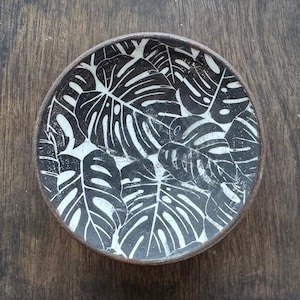 May include: A black and white ceramic plate with a monstera leaf pattern. The plate is round and has a slightly raised rim.