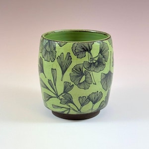 Puede incluir: Un vaso de cerámica con esmalte verde claro y base marrón oscuro. El exterior presenta un patrón repetitivo de hojas de ginkgo dibujadas con líneas negras. El interior está esmaltado en verde claro sólido y la superficie muestra una textura craquelada.